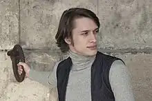 Un homme brun se tient devant un mur en pierre. Il tient avec sa main droite un anneau accroché au mur. Il est vétu d’un col roulé gris avec un gilet noir par dessus. Sa tête est tourné vers la droite.