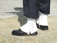 Guêtres d’apparat (blanches, à lacets blancs, évasées) en gros plan sur chaussures noires et pantalon noir&nbsp;; les ombres du soldat et de son arme sont visible sur le gravier.