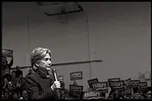 photographie : Hilary Clinton au micro dans une salle de sport, son nom est en arrière plan sur des pancartes